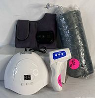 Photo of a white UV nail lamp with digital display, a white and blue digital infrared thermometer, a black heating pad rolled in plastic, and a grey heated knee brace with digital remote.
