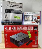 Boxed Full HD home theater projector by RCA, ProScan HDMI DVD player box, and Sylvania 7 inch WiFi digital photo frame box stacked together.