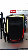 Front view of Ion Total PA Freedom speaker with yellow and red LED lights lit, next to folded black speaker stand and wireless microphone on table.
