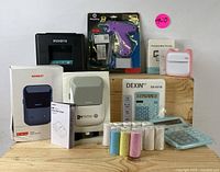 Overall set comprising NIIMBOT label printer with box and manual, purple glue gun in original packaging, DEXIN digital calculator with box, and multiple rolls of adhesive labels in various colors arranged on plywood surface.