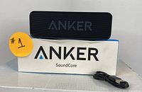 Black Anker SoundCore Bluetooth speaker atop original Anker SoundCore box with USB charging cable laid beside.
