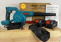 Cordless leaf blower laid on wooden surface with two batteries, charger, earplugs, cloth debris bag, original box and manual visible.