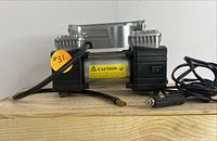 Side view of portable air compressor showing metal handle, black attached hose, power cord with car adapter, and caution label.