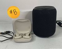 Photo showing black fabric smart Bluetooth speaker, white wireless earbuds in open charging case, and a USB charging cable on a white surface with orange sticker #8.