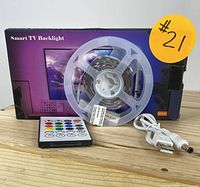 Photo showing the LED backlight strip on a plastic spool, remote control with color buttons, USB cable, and product box labeled Smart TV Backlight