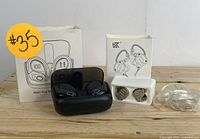 Two sets of wireless earbuds with their charging cases, original boxes, additional ear tips, and a clear wired earpiece arranged on a wooden surface.