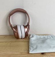 Front view of wireless over-ear headphones standing upright with white cushioned ear pads and folded design next to white protective carrying pouch.