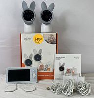 Two Aoneti baby cameras with bunny ears on top of the original box, plus the monitor, charging bases, cables, and manual arranged on a white surface.