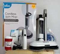 Full view of the cordless spin mop system including the box, mop with dual spinning pads, charging adapter, detachable handle and spray mechanism, user manual, and a cup for size reference.