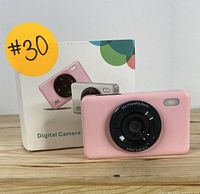 Front view of pink kid-friendly digital camera with lens and original boxed packaging in background