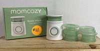 Momcozy bottle warmer box, warmer unit, and four green accessory inserts arranged on a wooden surface with product packaging in background.