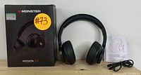 Monster Mission V1 wireless headphones placed on a wooden surface showing original packaging box, headphones, charging cable, and user manual arranged side-by-side.