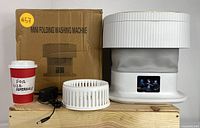 Portable mini folding washing machine with power adapter, washing basket, and a size reference cup placed beside the machine.