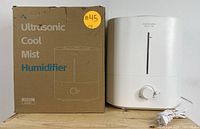 Asakuki Ultrasonic Cool Mist Humidifier placed next to its original cardboard box packaging, showing product branding and some basic text.