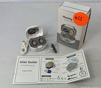 Photo of rechargeable hearing aids inside white charging case, USB cable, user guide, and cleaning tools on gray surface with product box behind.