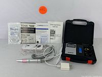TENS 7000 unit, electric nail drill handpiece with pink grip, power adapter, electrode leads, manuals, and registration cards arranged on white background.