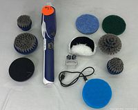 Handheld electric scrubber in blue and white with various round brush heads, several cleaning pads and a charging cable on a flat surface.