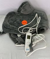 Folded grey plush heated shawl with attached white digital remote control and power cable placed on a white surface.