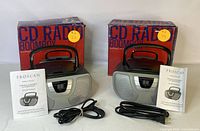 Two boxed Proscan CD radio boomboxes displayed with power cords and user manuals in front, showing original packaging and included items.