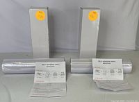 Two white self-adhesive vinyl rolls in plastic wrapping, each accompanied by a white box and a printed instruction sheet placed in front.