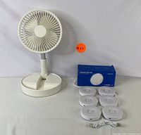 Front view of the white adjustable desk fan beside six white LED puck lights, USB cable, and the blue retail box for the puck lights.