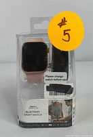 Proscan Bluetooth Smartwatch visible inside original plastic packaging with rose gold band attached and black band behind.