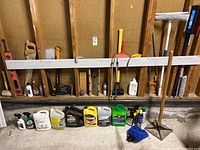 Garage wall with various tools hung or leaned including saws, axes, spirit levels, and a hoe. Containers of automotive fluids and sprays lined up on the floor.