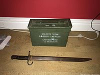 View of green metal ammunition box and antique bayonet with metal scabbard placed on floor against red wall