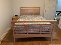 View of the bed frame with wooden headboard, footboard, mattress with floral cover, and side table beside the bed.