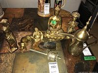 Seven brass items displayed on a tile surface including the seated Buddha, coffee pot, incense burner, baby shoe decor, elephant figurine, and standing female figurine taken from above at an angle.