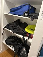Overall shelf showing various bags and backpacks stacked, including a blue Samsonite wheeled bag on top shelf, and black and red duffle bags beneath with a yellow neck support cushion visible.