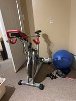 Photo of Kettler stationary bike with red handlebars and blue exercise ball, dumbbells and ankle weights near the wall in basement