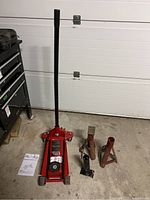 Photo showing the red Big Red hydraulic floor jack, black handle, two red metal jack stands on concrete floor, and instruction manual.