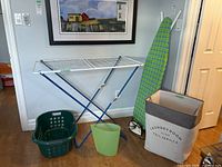 Metal and wire clothes drying rack, plastic laundry basket, fabric laundry hamper, ironing board, and plastic bucket arranged against a wall.
