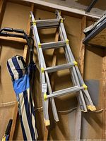 Front angle view of folded aluminum ladder with yellow locking knobs and protective feet, hanging in a garage next to a folded beach chair.