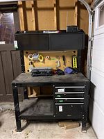 Front view of black steel workbench showing two-door upper cabinet, pegboard backing, work surface and lower shelf