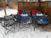 Outdoor shot of three chairs covered partially with snow, two blue and gray swivel chairs and one striped blue folding chair.