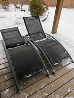 Two black plastic mesh patio lounge chairs with curved metal frames shown on snowy wooden deck from front angle.