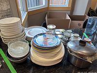 Overview of assorted plates, bowls, jars, and cups arranged on black granite counter beneath large windows.