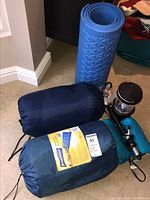 Blue foam sleeping mat rolled up, two sleeping bags in blue sacks, Coleman lantern, curling iron on floor.