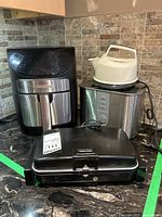 Black and stainless steel Gourmia air fryer, white kettle on black base, Delonghi toaster visible behind, black sandwich maker in front