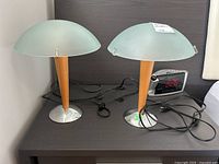 Two table lamps together on a dark surface showing wood bases, metal feet, frosted glass shades and cords stored loosely.