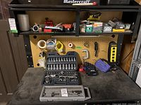 Full view of workshop table with various tools and hardware, two bit sets in front, Mastercraft socket wrench set open on table below workbench with pegboard.