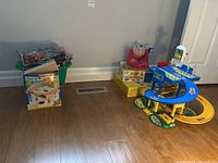 Wide shot showing wooden railway train set box and pieces beside various other toys including bingo game, parking garage, and green bin of toys.
