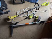 Wide view of Ryobi cordless trimmer, water sprayer handle, and Fiskars weeder laid on floor.