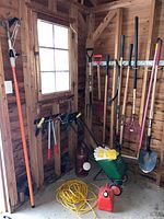 Photo of yard tools including long-handled shovels, rakes, seed spreader, gloves, and fuel container stored inside a wooden shed.