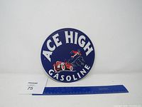 Full view of Ace High Gasoline metal round sign with a ruler for size reference, showing blue background, white text and red racecar and aviator graphic.