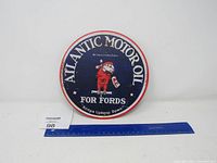 Photo showing the front of a round metal sign for Atlantic Motor Oil featuring a red parrot and text for Fords on a blue background with a red border.
