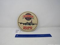 Front view of the metal round sign showing the bomber airplane graphic with a ruler for scale.