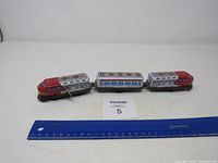 Full view of the three connected cars of the metal wind-up toy train against a white background with a blue ruler for scale.